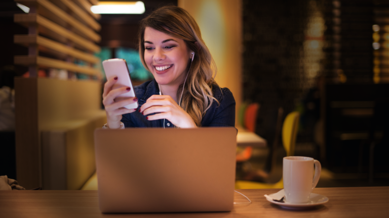 Redes Sociais - Mulher sentada à mesa de um café em frente ao seu laptop e a uma xícara enquanto segura o celular e o olha sorridente.